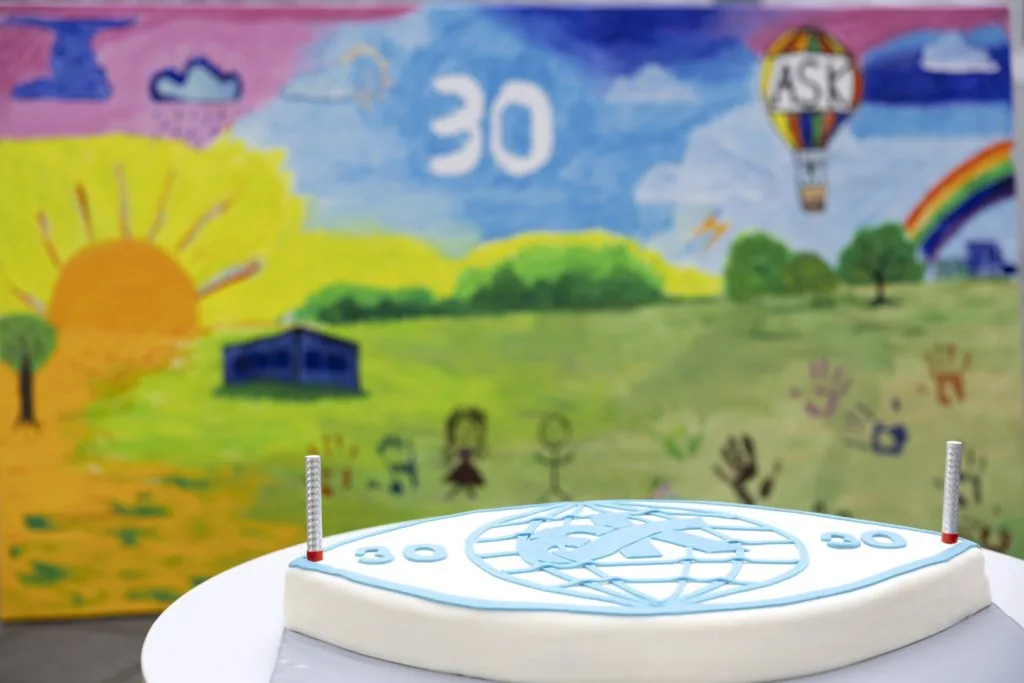 Colourful anniversary painting with a large “30”, the ask logo on a hot-air balloon and a rainbow, behind a cake decorated with the ask logo